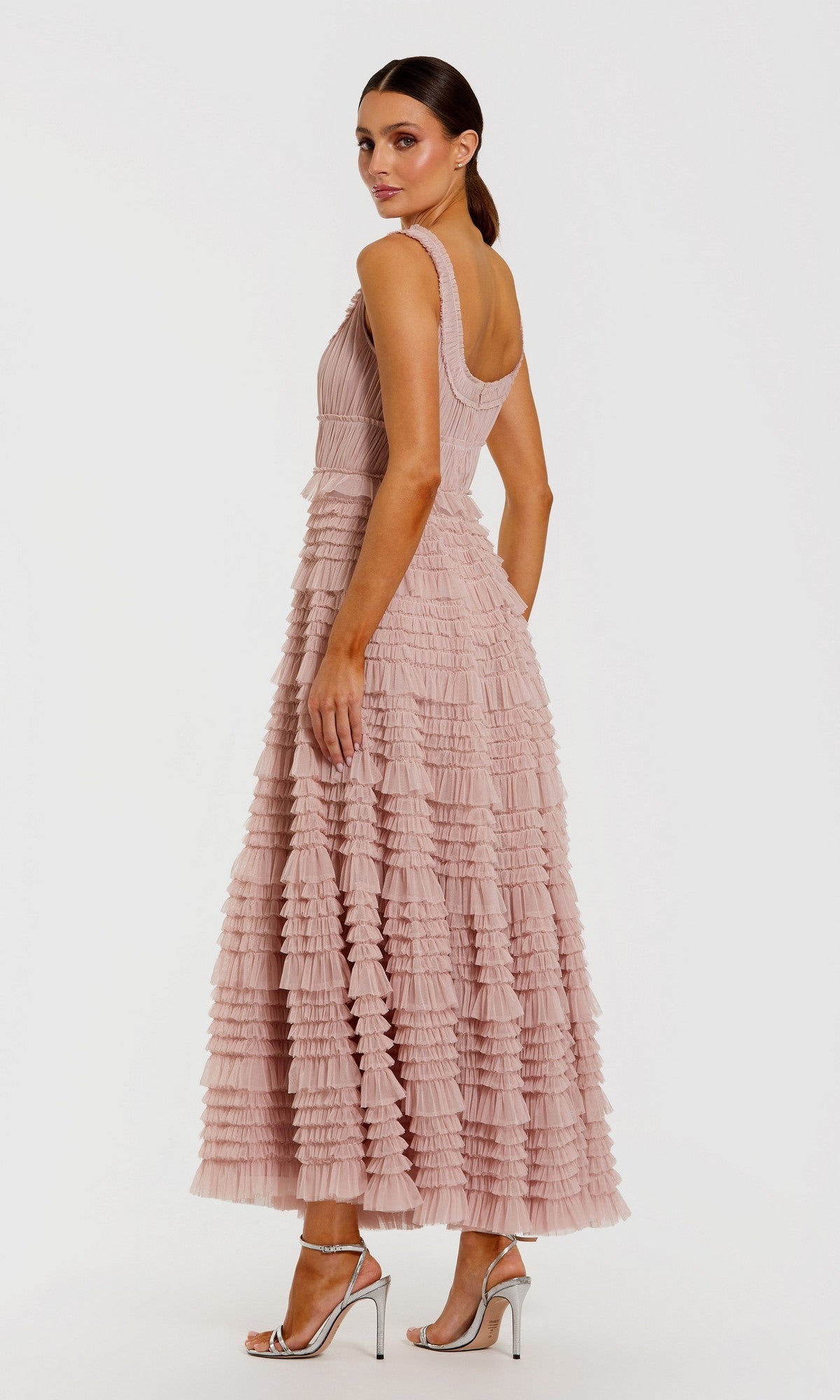 A woman stands in the Tea-Length Party Dress: Mac Duggal 8292—a sleeveless, blush pink mesh midi cocktail dress featuring delicate ruffle detailing and an elegant A-line tiered skirt—paired with silver high-heeled sandals, facing slightly away against a plain white background.